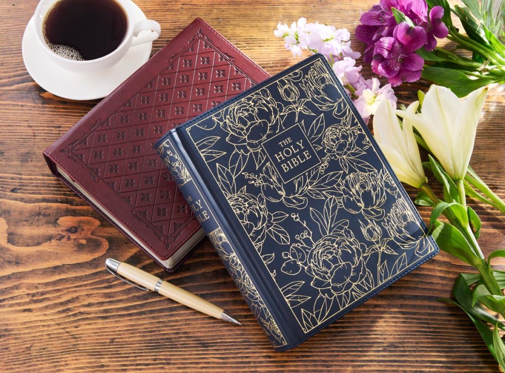 Burgundy with diamond pattern journaling Bible underneath navy blue floral pattern journaling Bible on wooden table with coffee cup and flowers