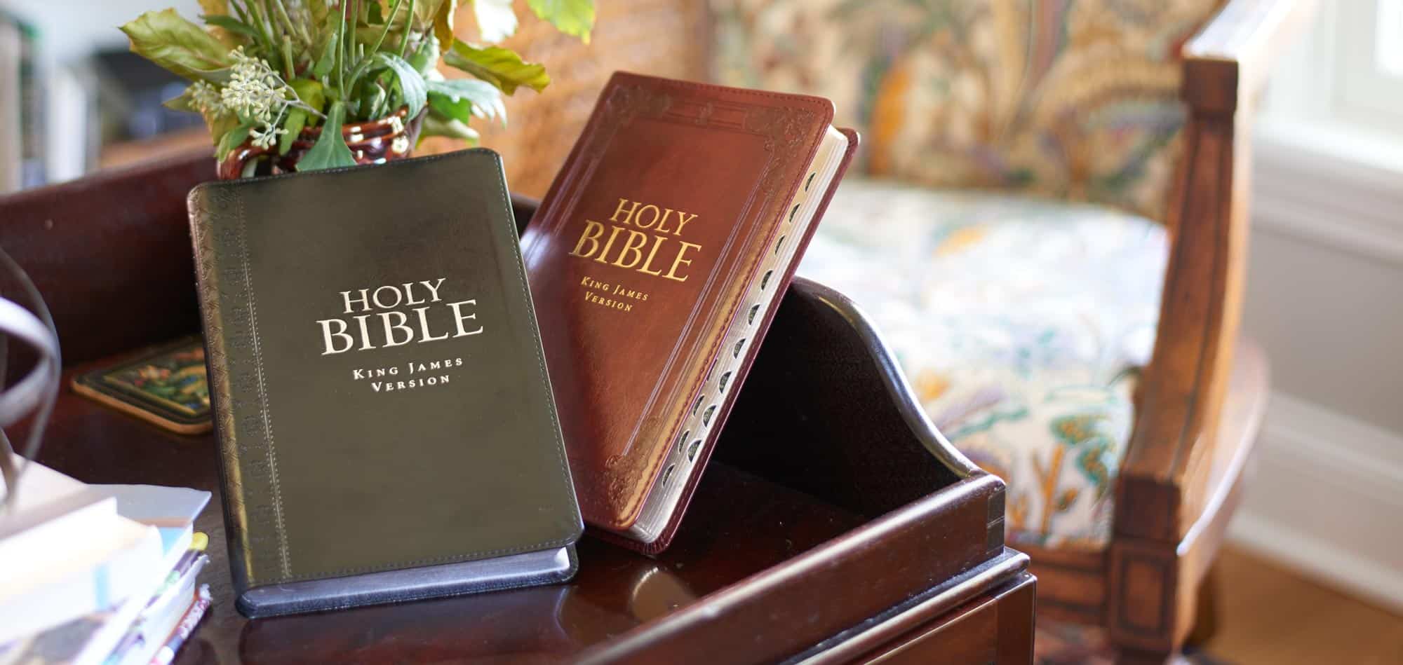 Two Bibles, one black and one brown, on a wooden table with a vase of greenery and chair in the blurred background